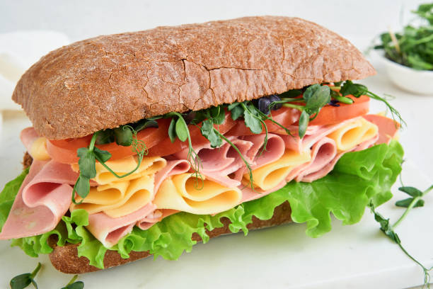 Sandwich. One fresh big submarine sandwich with ham, cheese, lettuce, tomatoes and microgreens on light background. Healthy breakfast theme concept, school lunch, breakfast or snack.