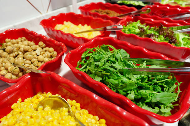 A variety of fresh salad ingredients are neatly presented in red dishes forming a vibrant buffet line. The bowls are filled with crisp greens, sweetcorn, and other tasty vegetables, inviting diners to create their own salad masterpiece. Each container is equipped with serving tongs or spoons for convenience, and condiments are provided along the table to enhance the flavors.