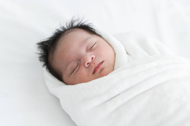 istock Adorable newborn baby wrapped in white swaddle towel lying on bed and sleeping peacefully. Sleeping cute infant in a wrap on white blanket. Beautiful close up portrait of little child girl 19 days 2148105321