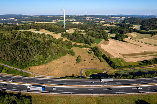 Aerial view of a rural highway with truck and car traffic at dusk. Wind turbines and a rolling landscape are visible in the background.