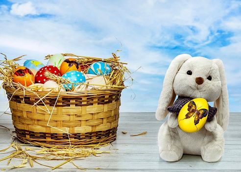 Easter eggs in a wicker basket on a white table and a toy rabbit with an egg close-up against the blue sky.