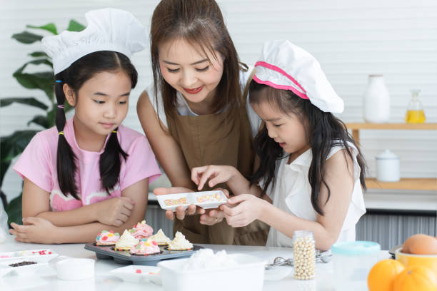 istock Mother and two daughters with chef hat and apron enjoy baking, decorating cupcake with jelly and icing sugar at kitchen at home together. Little children have fun with mom. Happy Asian family 2148110381
