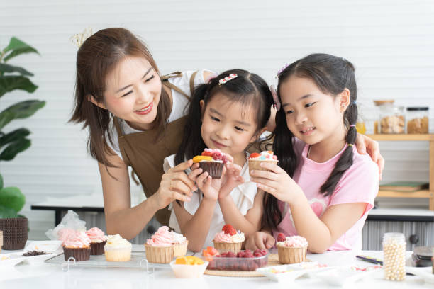 istock Mother and two daughters enjoy making, decorating cupcake with fresh fruits and icing sugar at kitchen at home together. Children have fun with mom. Happy Asian family 2148110392