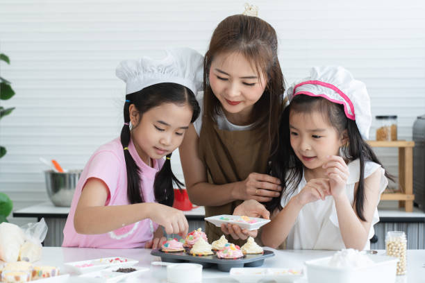 istock Mother and two daughters with chef hat and apron enjoy baking, decorating cupcake with jelly and icing sugar at kitchen at home together. Little children have fun with mom. Happy Asian family 2148110457