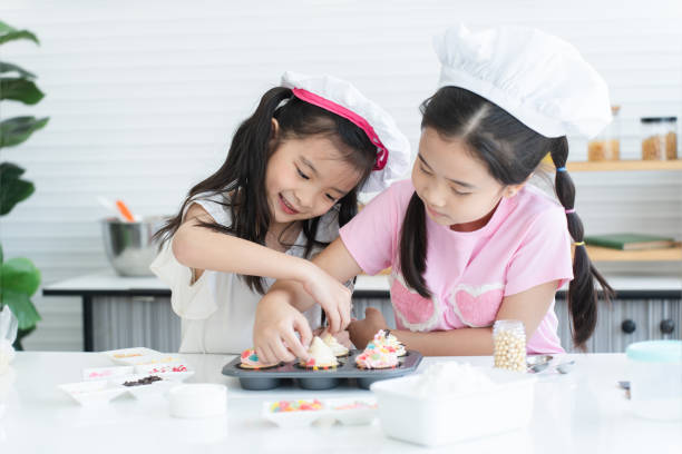 istock Two cute sisters with chef hat enjoy baking, decorating cupcake with jelly and icing sugar at kitchen at home. Little children girls have fun together. Happy Asian family 2148110537