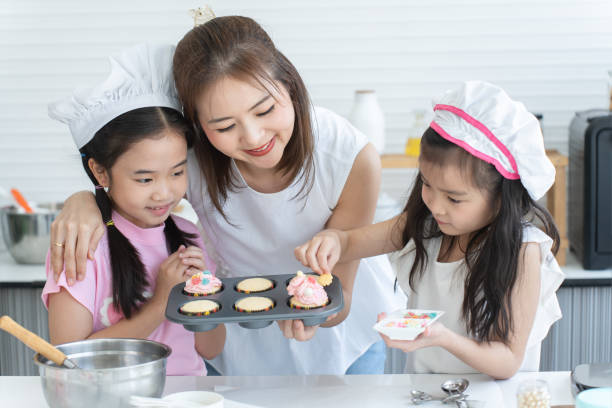 istock Mother and two daughters with chef hat enjoy baking, decorating cupcake with jelly and icing sugar at kitchen at home together. Little children have fun with mom. Happy Asian family 2148110549