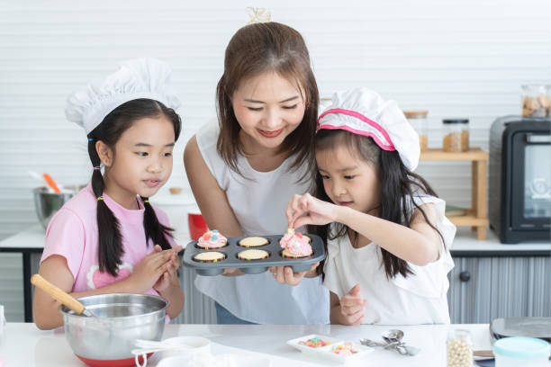 istock Mother and two daughters with chef hat enjoy baking, decorating cupcake with jelly and icing sugar at kitchen at home together. Little children have fun with mom. Happy Asian family 2148110566