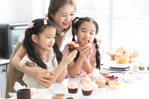 Mother and two daughters enjoy making, decorating cupcake with fresh fruits and icing sugar at kitchen at home together. Children have fun with mom. Happy Asian family. Selective focus on girl in pink