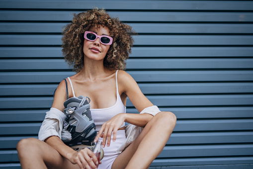 Trendy young woman with curly hair wearing sunglasses and roller skates rests by a blue wall