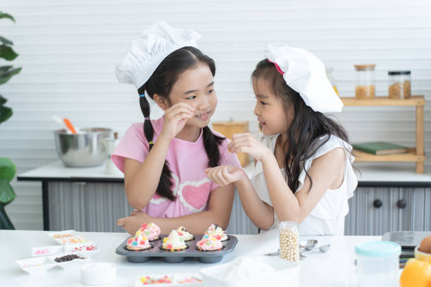 istock Two cute sisters with chef hat enjoy baking, decorating cupcake with jelly and icing sugar at kitchen at home. Little children girls have fun together. Happy Asian family 2148110599