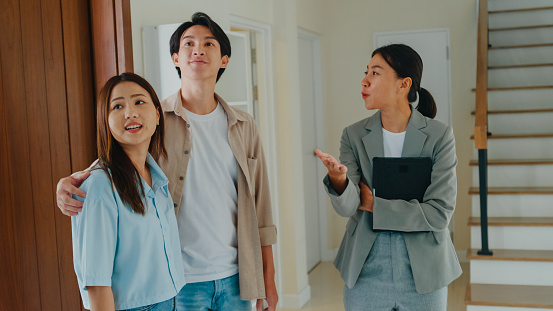 Asian female real estate agent explaining and showing new houses to tenants for rent or buy. Landlord selling real estate to happiness couple customer for investment concept.