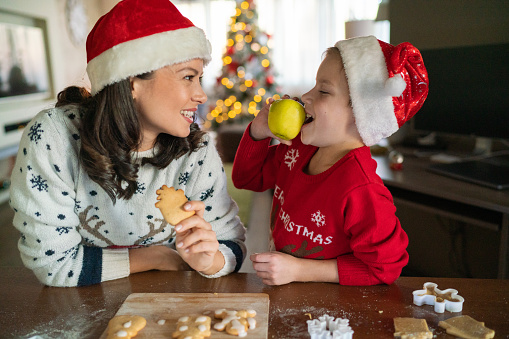 Happy little boy eating an apple while making  Christmas cookies whit his mother