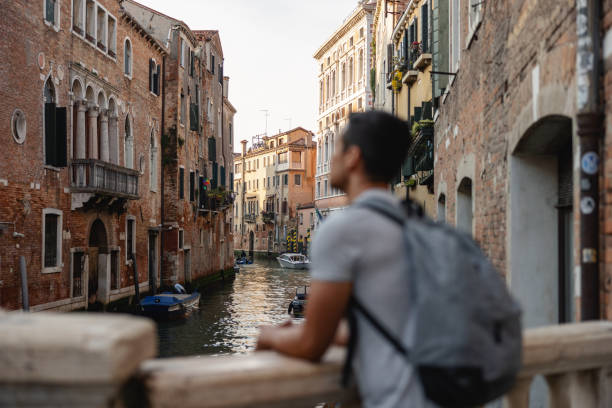 Back view of a single male tourist with a backpack admiring a picturesque canal in Venice, Italy during travel.