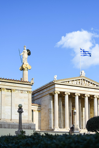 Iconic statue of Athena stands tall beside the Greek flag fluttering proudly in the wind.