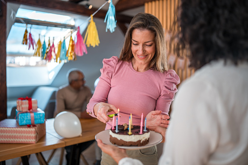 A middle-aged Caucasian female lighting candles on a chocolate birthday cake, smiling at a diverse family gathered in a home decorated with balloons and party accessories.