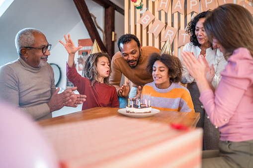 A diverse, multigenerational family with mixed ethnicities celebrates a young female's birthday. The group, happily engaged, focuses on the birthday cake as they prepare to blow out the candles in a home setting.
