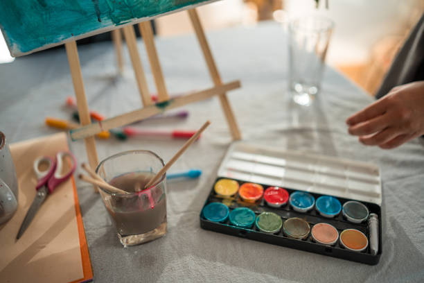 A close-up view of art supplies including watercolors, brushes, and scissors prepared on a table with an easel and blank canvas, ready for a creative painting session.
