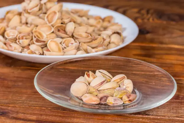 Partly peeled roasted salted pistachio nuts on the glass saucer on a blurred background of the empty shells on a big dish on a rustic table, side view in selective focus
