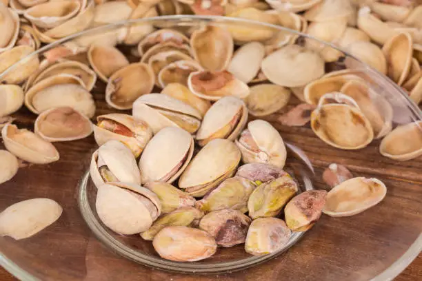 Partly peeled roasted salted pistachio nuts on the transparent glass saucer against the empty shells on a rustic table, close-up in selective focus