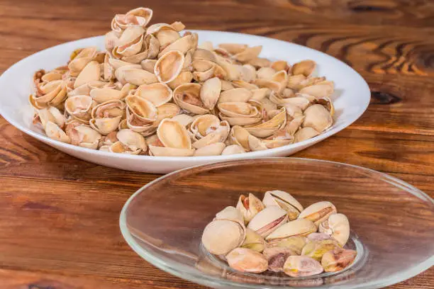 Partly peeled roasted salted pistachio nuts on the glass saucer on a background of the empty shells on a big dish on a rustic table, side view close-up