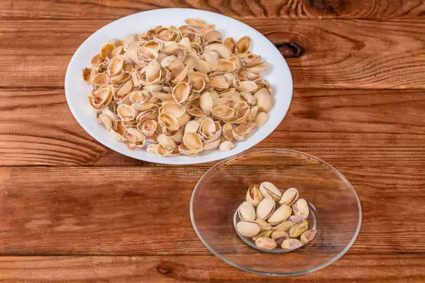 Partly peeled roasted salted pistachio nuts on the glass saucer against the empty shells on a big dish on a rustic table