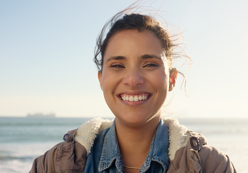 Woman, portrait and smile on beach with wind for travel, holiday or vacation outdoor in nature with relax. Tourist, happy and person with face in morning for fresh air, confidence and sky with breeze