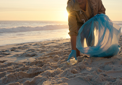 Beach cleaning, bottle and person volunteering for earth day, pollution and environment in sand and hands closeup. Recycling bag, plastic and garbage to stop pollution and climate change by the ocean