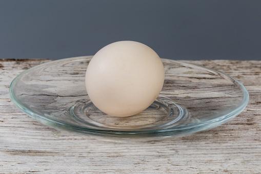 Whole boiled chicken egg in a white shell on glass transparent saucer on a old cracked wooden surface, side view in selective focus on a gray background