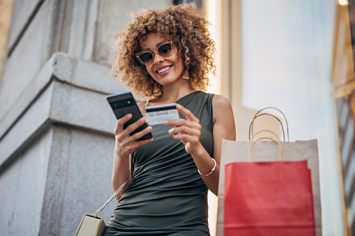 The young adult woman enjoying a joyful shopping spree with her smartphone and credit card on a sunny day in the urban fashion retail district