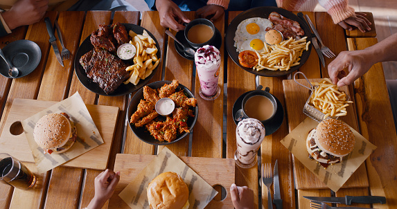 Top view, hands and fast food on table at restaurant for dinner or lunch, people with cuisine and catering. Burger, fries and milkshake drinks with wings for nutrition and eating meal for supper