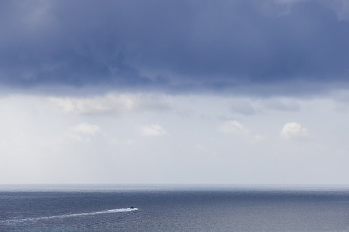 Under a canopy of gathering storm clouds, a lone speedboat ventures towards the distant horizon, contrasting the tranquil sea with nature's brewing intensity