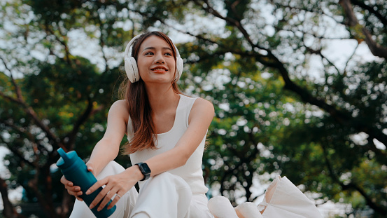 A woman in white enjoys her favorite melodies, sitting under the shade of trees with headphones and a water bottle, savoring a musical break