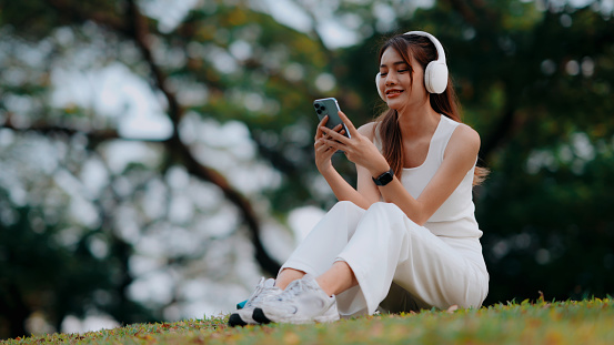 A young woman enjoys a peaceful moment in a lush park, engrossed in her music and smartphone, embodying relaxation and digital lifestyle. Ideal for promoting wellbeing apps or outdoor audio equipment.