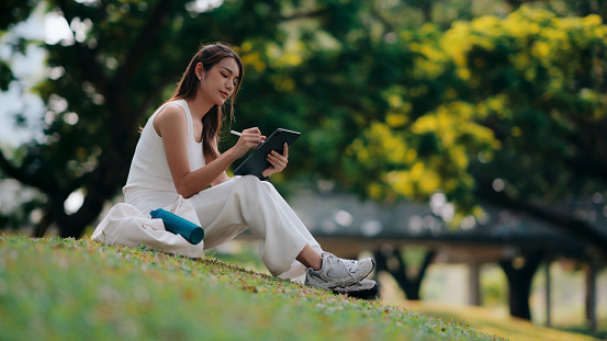 Amidst the serenity of greenery, a young woman sits cross-legged on the grass, focused on her tablet, harmonizing digital life with nature's calm