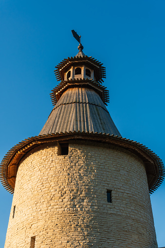 A powerful round tower of the protective wall of the old city with a wooden roof and an observation post on top, a protective structure with a hipped roof, a weather vane, bright rays of the morning sun, city landscape, history and culture, dawn, autumn.