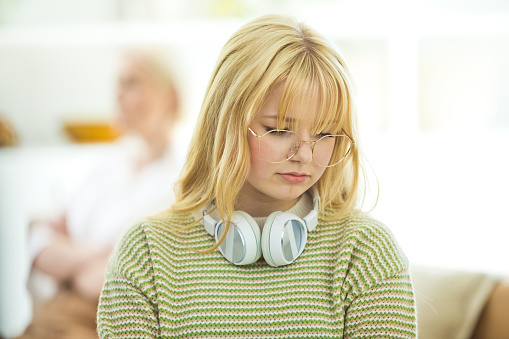 Worried teenage girl wearing headphone sitting on sofa in the living room at home with her defocused mother in the background. They are in conflict.