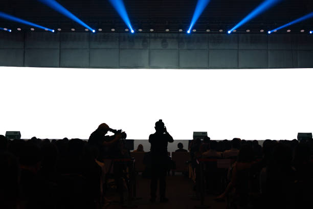 istock Front view of mockup on big blank white screen with space in empty huge hall with scene and silhouette of audiences 2148134457