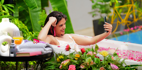Happy Indian young adult unwinding girl sitting in bathtub fresh morning bathing in rose petals water enjoying summer vacation at hotel resort. Beautiful smiling woman distant talking using mobile phone outdoor home
