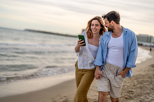 Couple enjoy sunny day on the beach, they are looking at photos on smart phone.