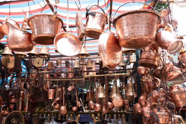 A close-up shot of a line of traditional, antique copper pitchers, urns, or coffee pots sitting on a polished wooden surface. The highly reflective copper material catches the light, showing a warm, reddish-brown metallic texture. The composition uses selective focus (shallow depth of field) to highlight the pot closest to the viewer while the rest blur softly into the background, creating a visual rhythm and emphasizing craftsmanship and traditional kitchenware.