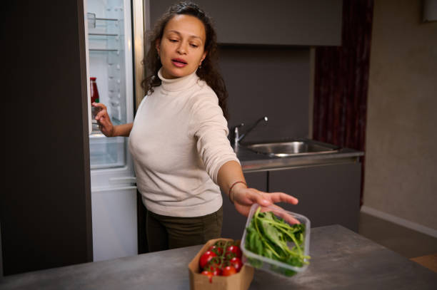 istock Young woman standing by fridge, pulling out a cardboard crate with fresh spinach leaves, putting it on the table near a carton box of tomatoes cherry, preparing healthy dieting salad at home kitchen 2148137211