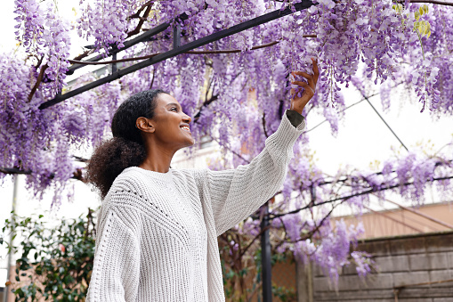 A cheerful black African American woman is in the backyard holding the purple wisteria flower in the square metal garden arch