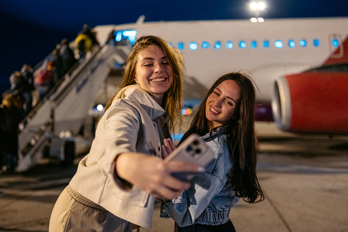 Two female friends taking selfies using a smart phone while boarding an airplane at night in Spain.