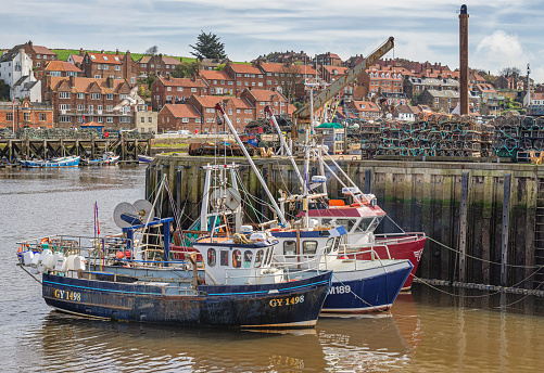Whitby, UK.  March 31, 2024.  Three small fishing boats are moored alongside a quay and buildings are in the background. Lobster pots are stacked on the quayside.