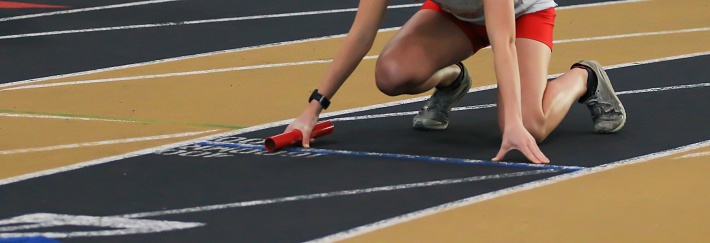 Female runner in the on your mark position holding a baton on an indoor track