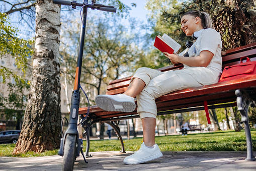 Woman enjoying on sunny day in public park reading on park bench near her electric scooter