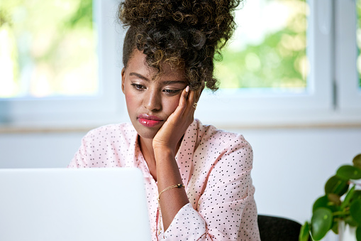 Worried afro american young woman wearing pink shirt sitting at the desk at home and using laptop. Home office concept.