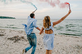 Happy married couple holds in hands smoke bomb. Baby shower. Twins: boy and girl. Back view. Man and woman having fun running on sand and holding colorful blue and red or pink smoke bombs on the beach