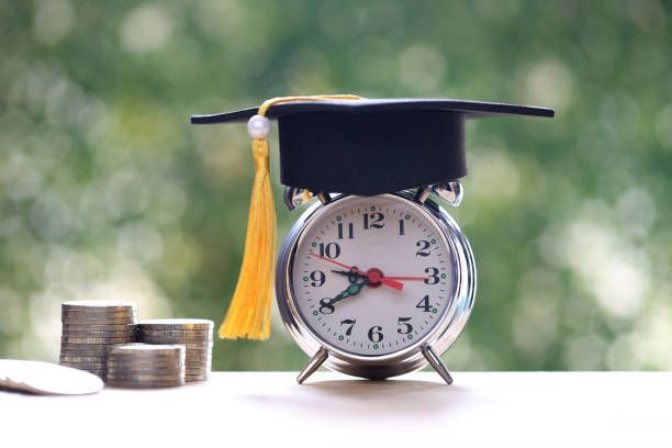istock Graduation hat on alarm clock with stack of coins money on natural green background, Saving money for education concept 2148144845