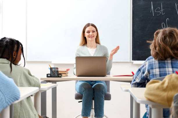 istock Smiling teacher talking to pupils while sitting at the desk in classroom, using laptop 2148145287